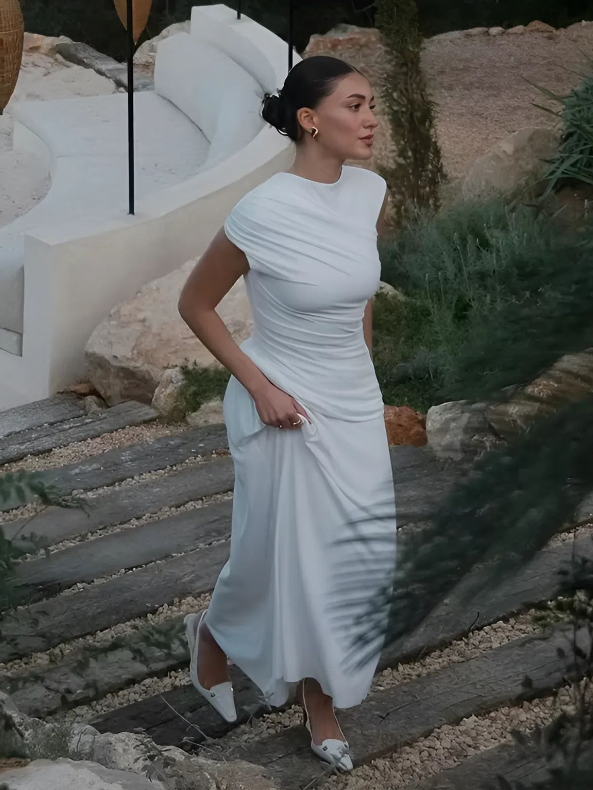 Stylish white bodycon maxi dress with asymmetric pleats, photographed on stairs for a classy look.